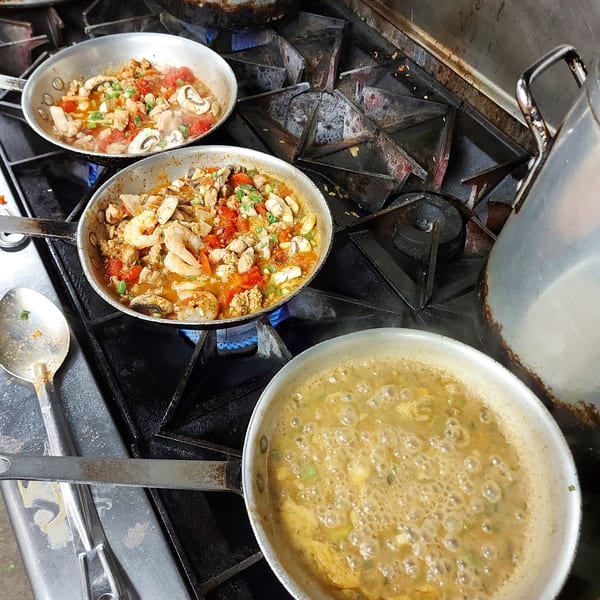 Three pans of Southern food, two with shrimp and vegetables, one with a bubbling sauce, cooking on a stove.